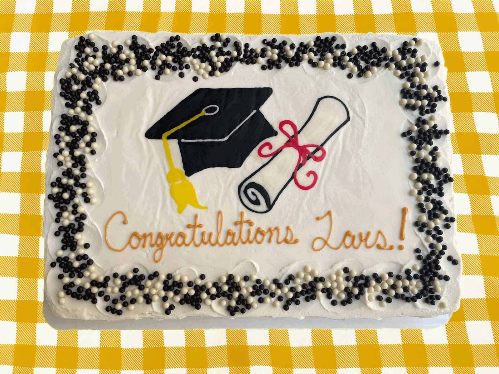 A rectangular white graduation cake decorated with a black graduation cap, a diploma tied with a red ribbon, and the words "Congratulations Lars!" in orange cursive, all framed by a border of black and white spherical sprinkles, set on a yellow and white checkered tablecloth.