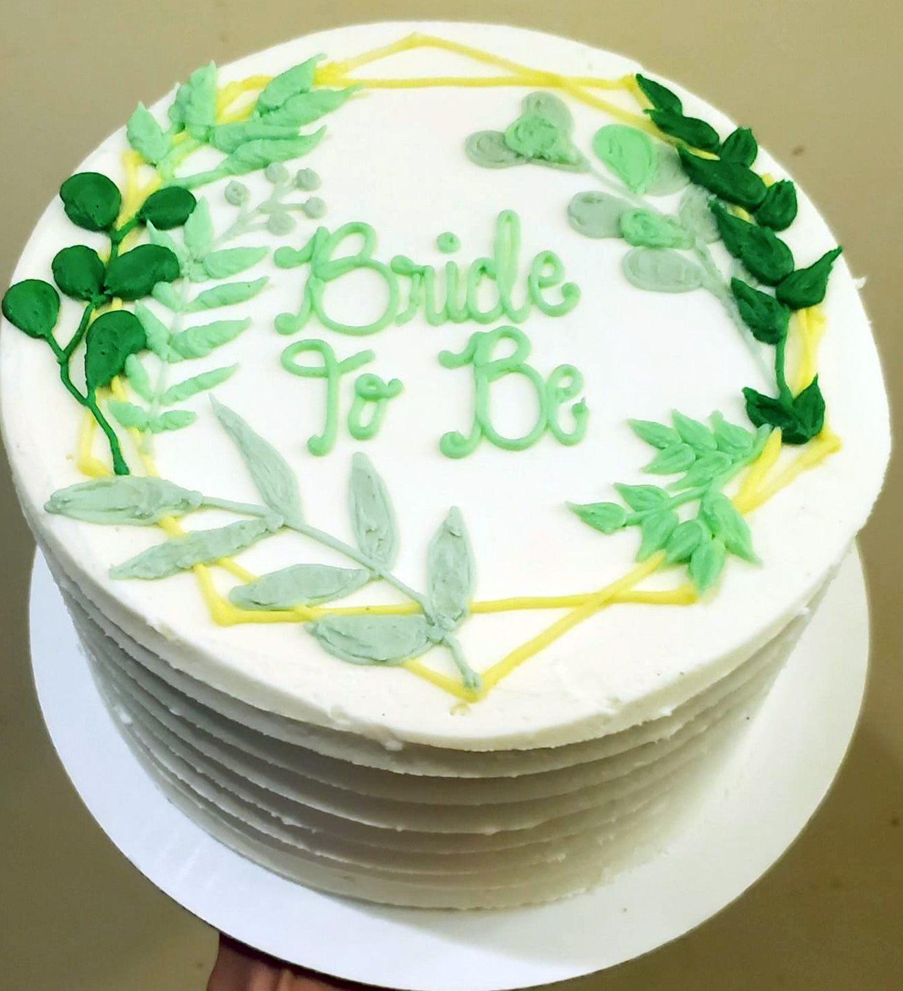 A round white cake decorated with green iced leaves, yellow branches and green 'bride to be' text.
