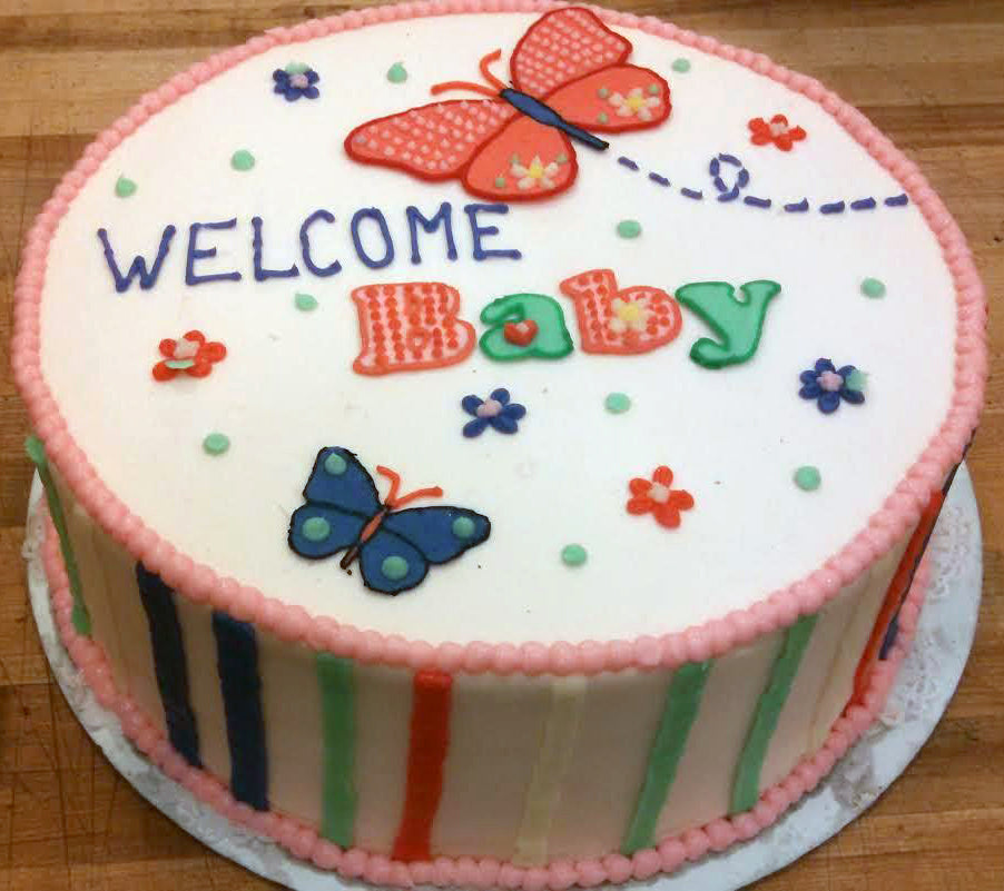 A white round cake decorated with colorful butterflies and flowers, featuring the words 'WELCOME Baby' in blue, red, and green frosting. The sides of the cake have vertical stripes in blue, green, and red.