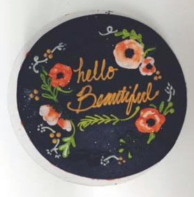 A round, dark-colored cake with the words "hello Beautiful" written in a stylized, golden script. The text is surrounded by illustrations of red and orange flowers with green leaves and white flourishes. 