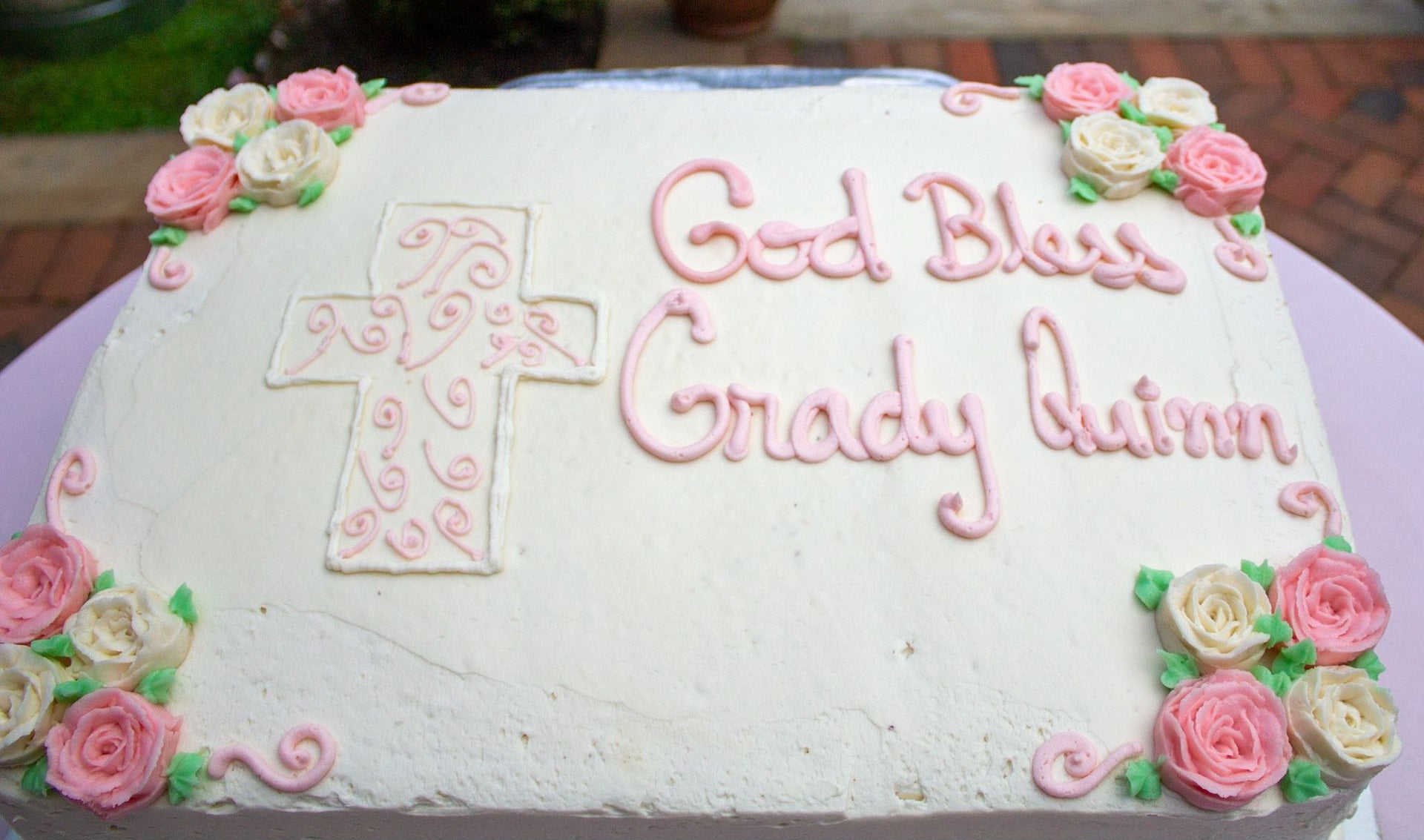 A large white rectangular cake decorated with pink and white frosting roses, a pink piped cross on the left, and the words 'God Bless Grady Quinn' in pink script on the right.