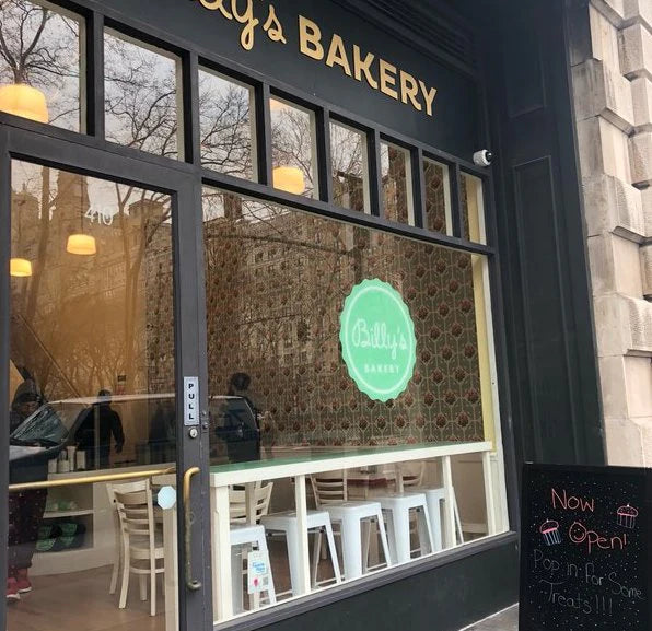 Exterior view of Billy's Bakery storefront, featuring a prominent green and white logo, reflections of trees and buildings in the windows, and a 'Now Open!' sign on the right.