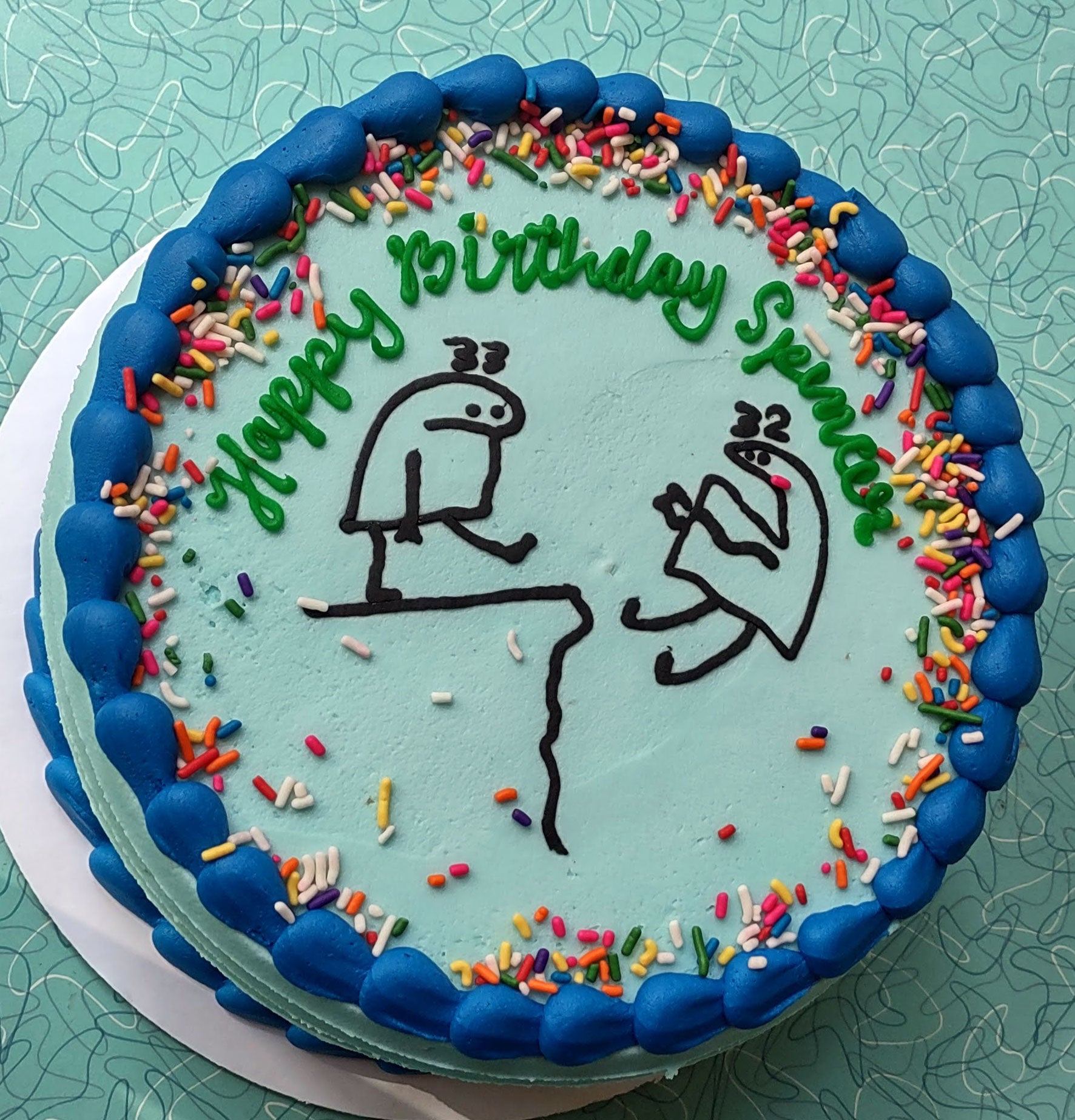 A light blue birthday cake with blue piped frosting and colorful sprinkles. The cake is decorated with green icing that reads "Happy Birthday Spencer" and features two black outline stick figures, one sitting on a cliff edge and another floating in the air.