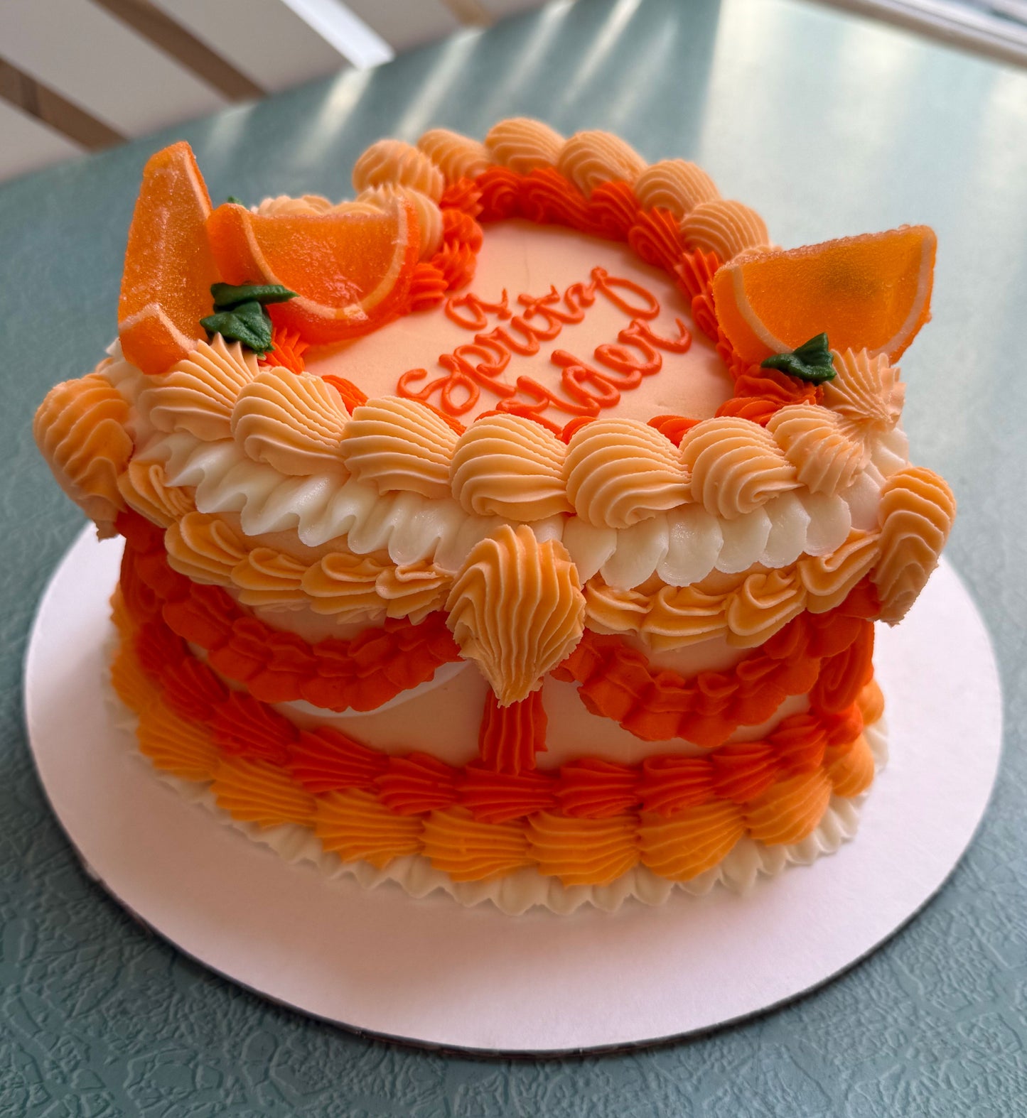 decorated cake with orange buttercream frosting and candied orange wedges, with the text 'A Spritz Older' written on top.
