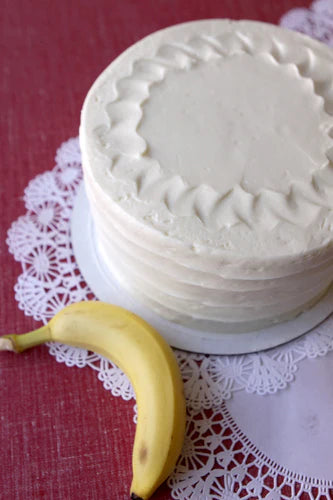 A whole banana cake with white frosting on a white lace doily, with a ripe yellow banana placed in front of it on a red surface.