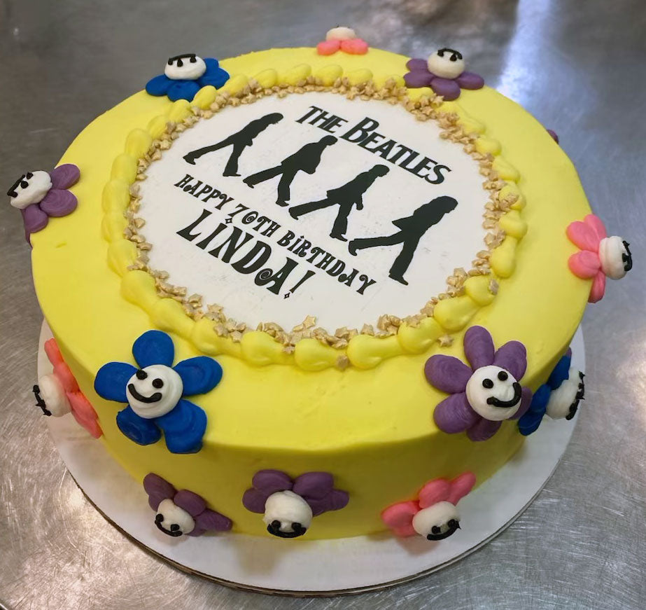 A vibrant yellow birthday cake decorated with The Beatles Abbey Road silhouette, text "HAPPY 70TH BIRTHDAY LINDA!", and colorful flower-shaped decorations with smiling faces.