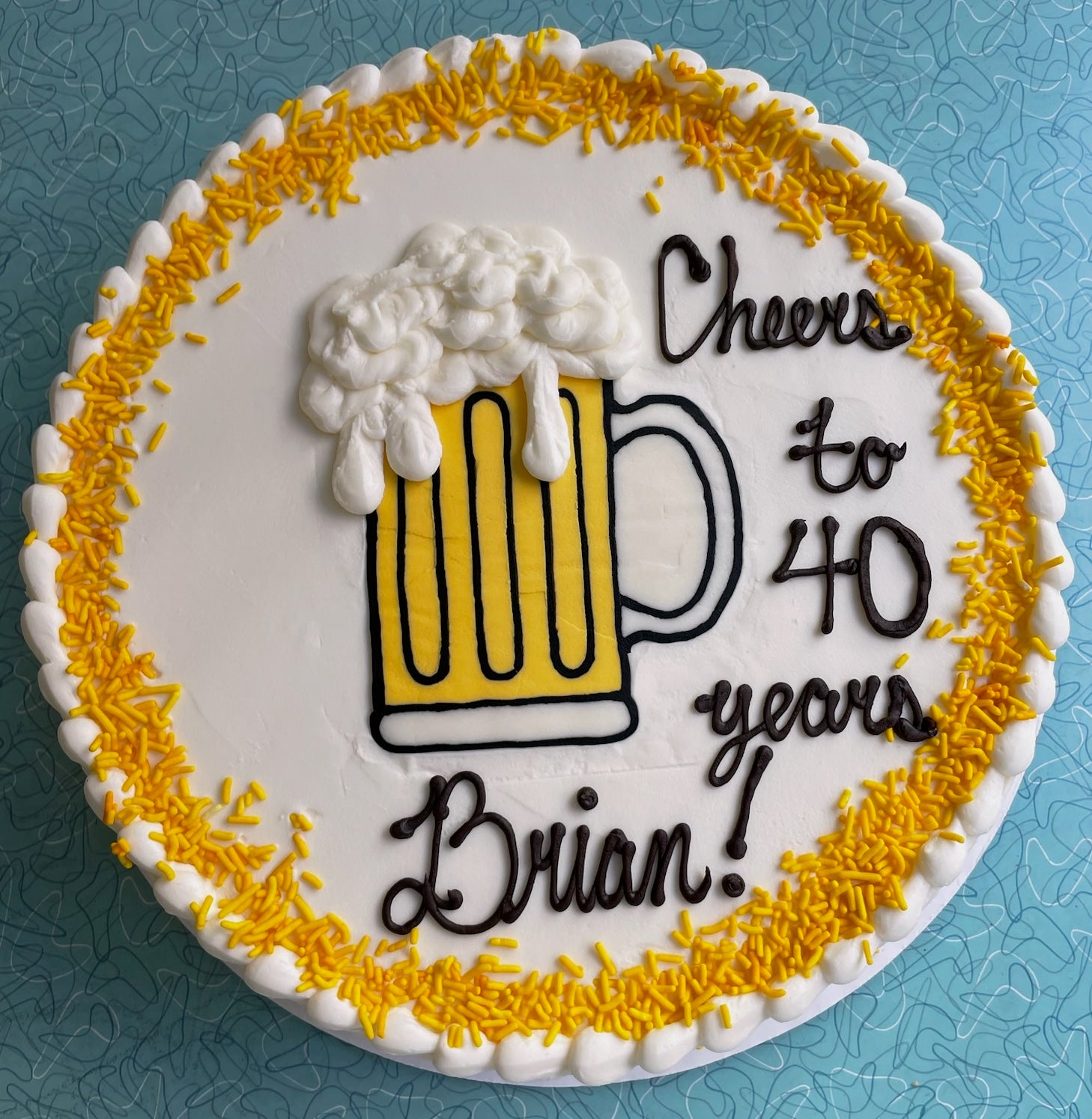Round white birthday cake with a beer mug design on a blue table, decorated with yellow sprinkles and black script text that reads 'Cheers to 40 years Brian!'