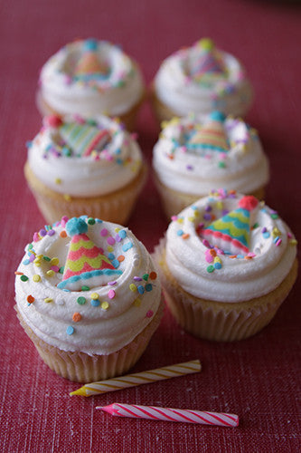 A set of cupcakes with white frosting and multicolored sprinkles on a red surface, with a birthday candle beside them. Candles NOT included. 