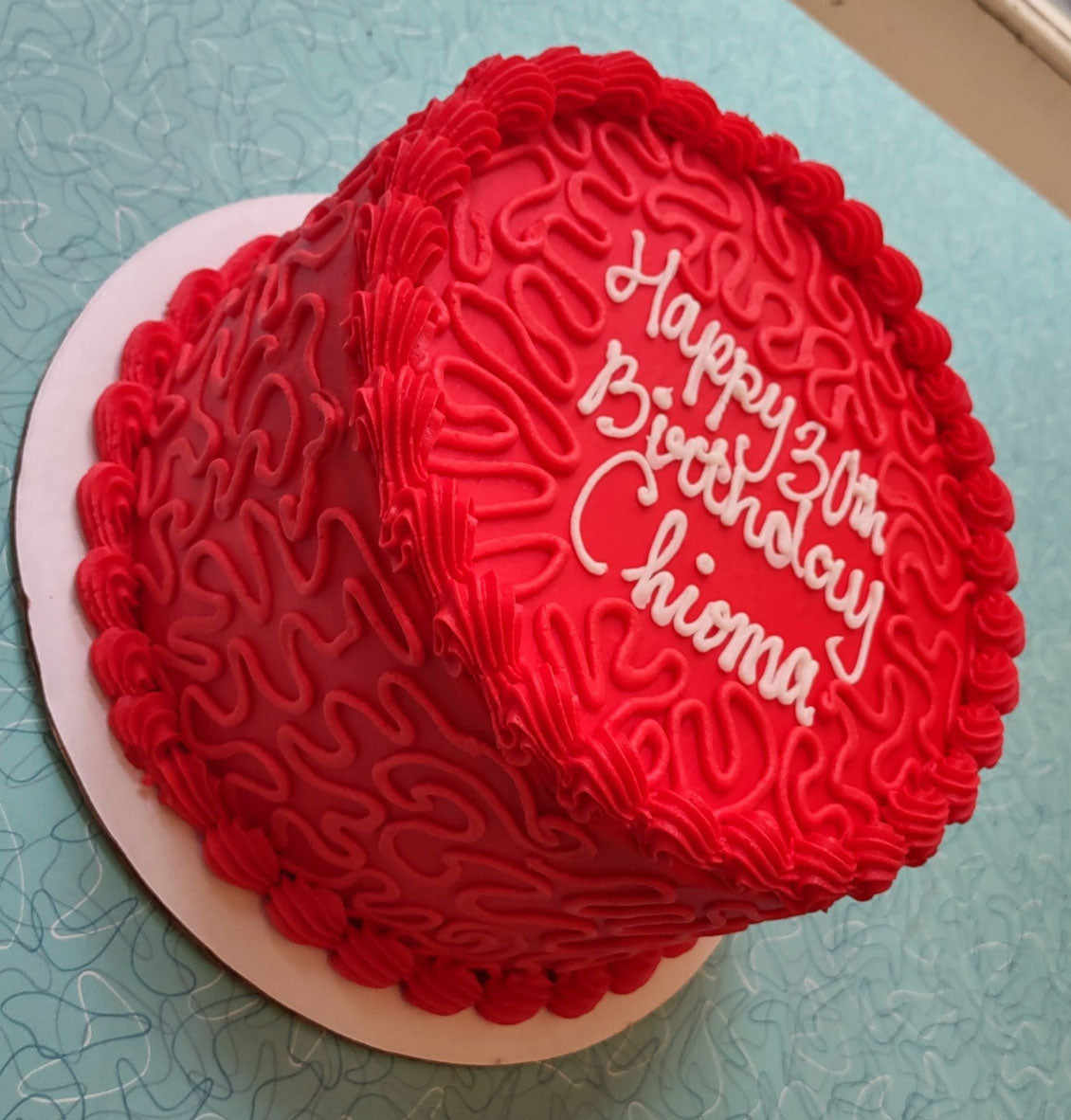 A vibrant red heart-shaped birthday cake with white frosting spelling "Happy 30th Birthday Chioma" on top, featuring intricate piped details around the edges and on the sides, resting on a white cake board.