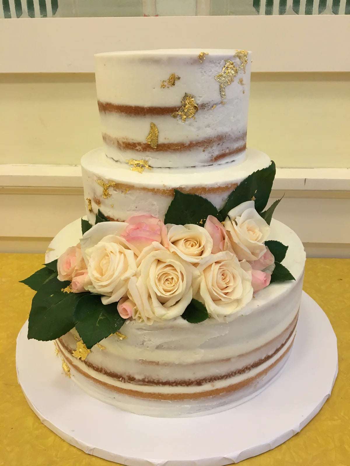 A two-tiered semi-naked wedding cake adorned with light pink and white roses, green foliage, and gold leaf accents.