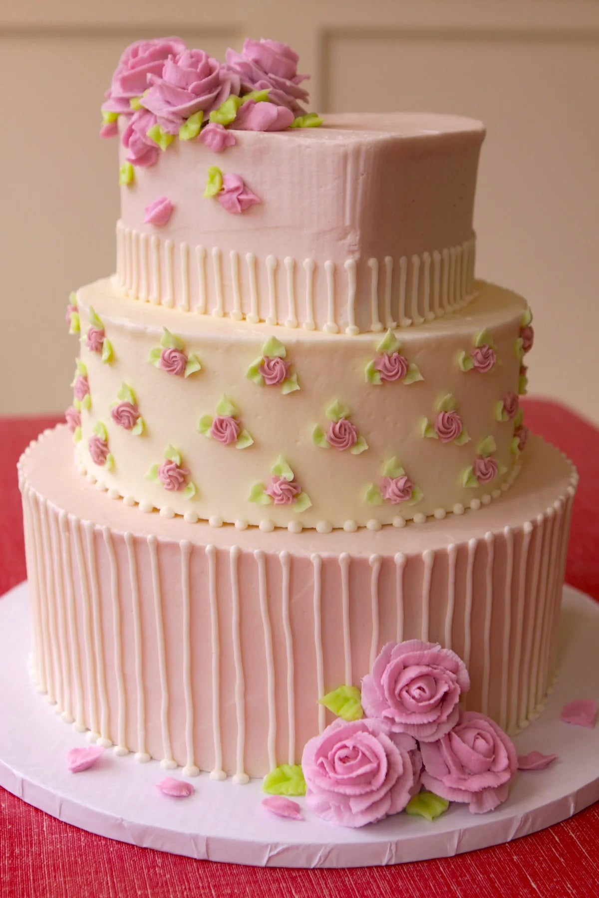 A three-tiered pink and white cake decorated with numerous pink roses and green leaves. The bottom tier features vertical piped lines, the middle tier has small piped rosebuds and stripes, and the top tier is smooth with a cluster of larger roses on top. More large pink roses are at the base of the cake.
