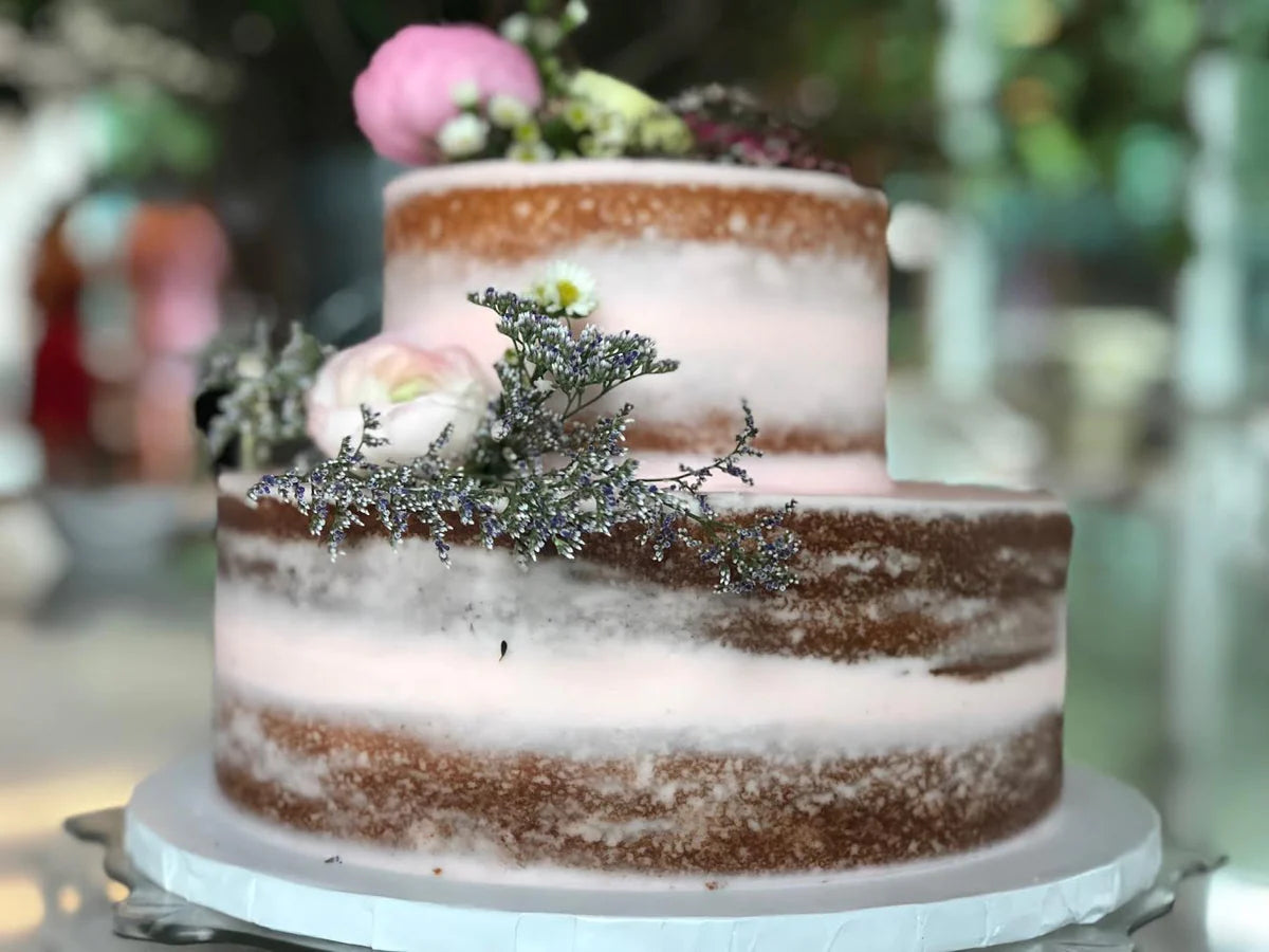 Two-tiered semi-naked wedding cake with light pink frosting, adorned with fresh flowers including white, pink and purple dried florals.