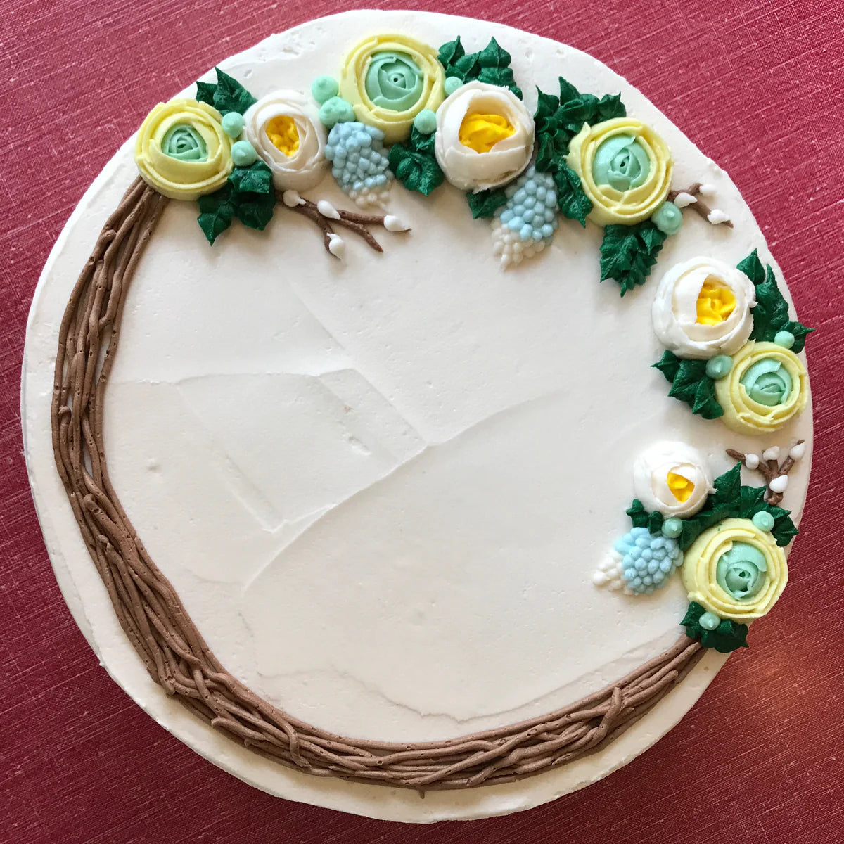 A white frosted cake decorated with a brown buttercream branch wreath on the lower left, adorned with yellow, white, and light blue buttercream flowers and green leaves on the upper right.