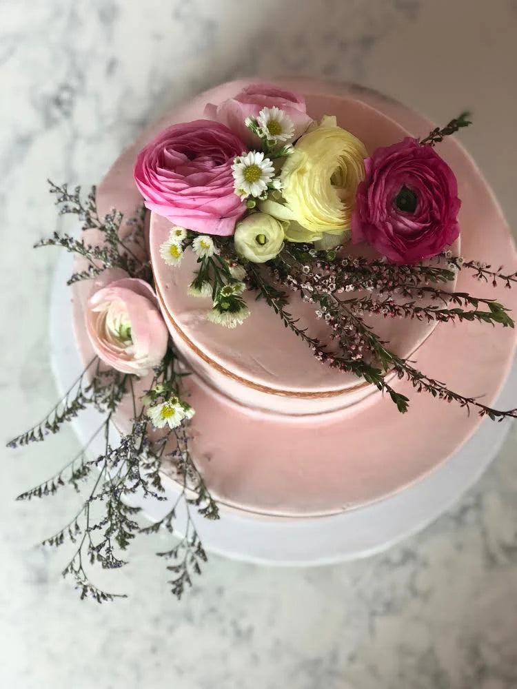 A two-tiered pink cake decorated with fresh flowers including pink and yellow ranunculus, small white daisies, and delicate green foliage.