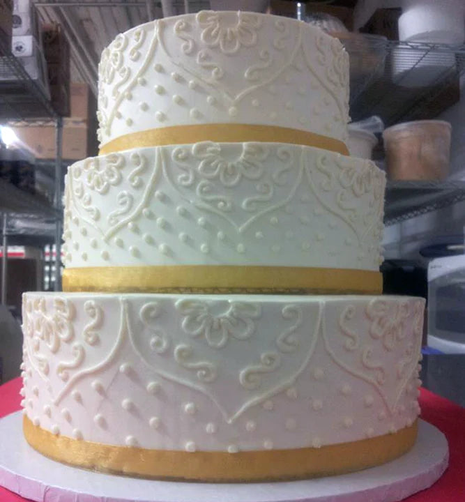 Three-tiered white wedding cake with intricate white buttercream scrolling and Swiss dot details, accented with gold ribbons on each tier.