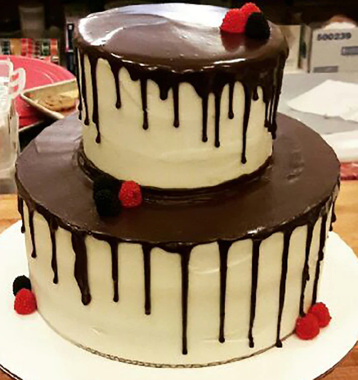 A two-tiered white cake with chocolate ganache drip, topped with fresh raspberries and blackberries.