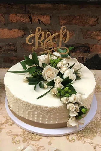 A white, single-tier wedding cake decorated with white roses, green foliage, and a gold "R & P" cake topper, set against a blurred brick background.