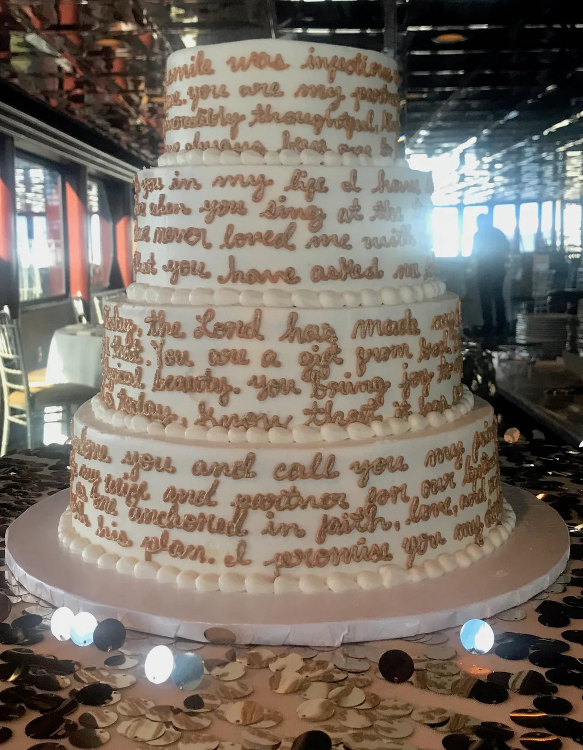 Four-tiered white wedding cake with gold script lettering, featuring personalized vows, on a table with scattered confetti and soft lighting.