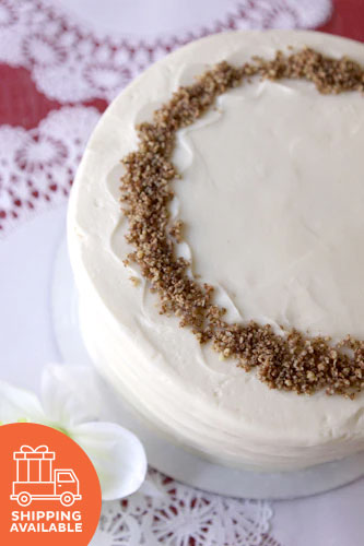 A round carrot cake with cream cheese frosting and chopped pecans on top, displayed on a white plate with a red and white lace background.