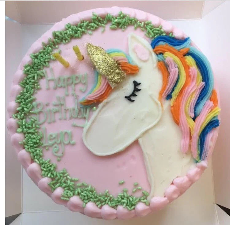 A round pink birthday cake with a white unicorn head, rainbow mane, and gold glitter horn. The cake is decorated with green sprinkles forming a wreath and pink frosting dots around the edge. "Happy Birthday Leya" is written on the cake in green frosting.