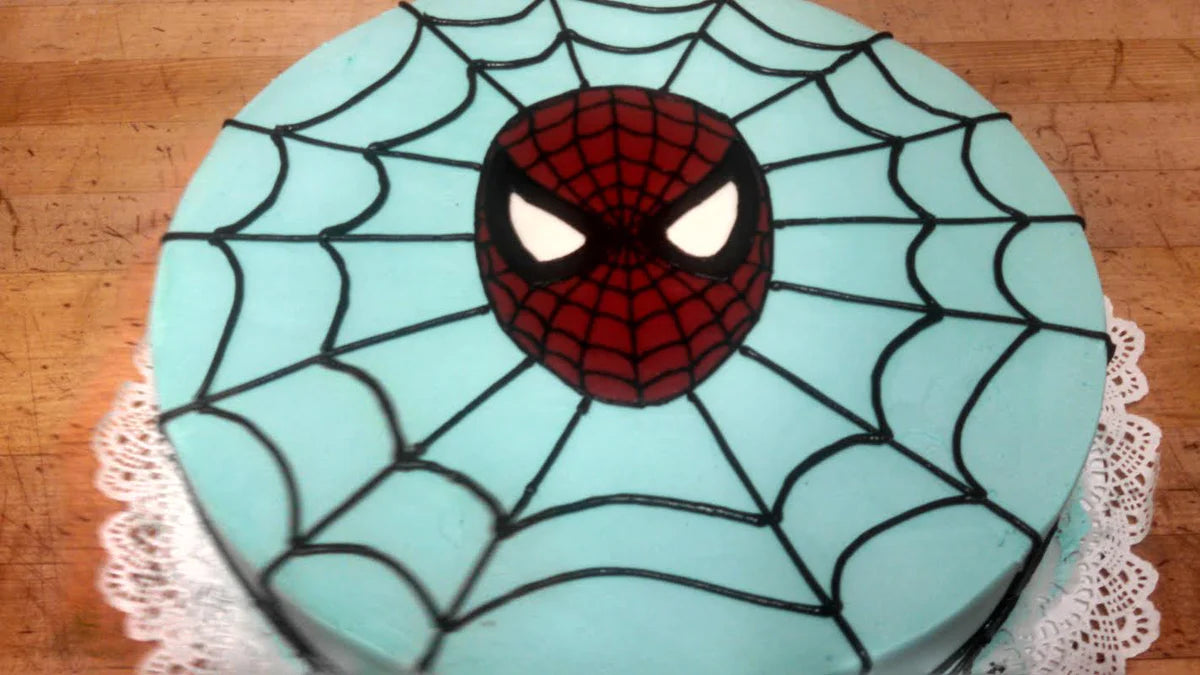 Round Spider-Man cake with light blue frosting, black spider web design, and a red and black Spider-Man mask in the center.