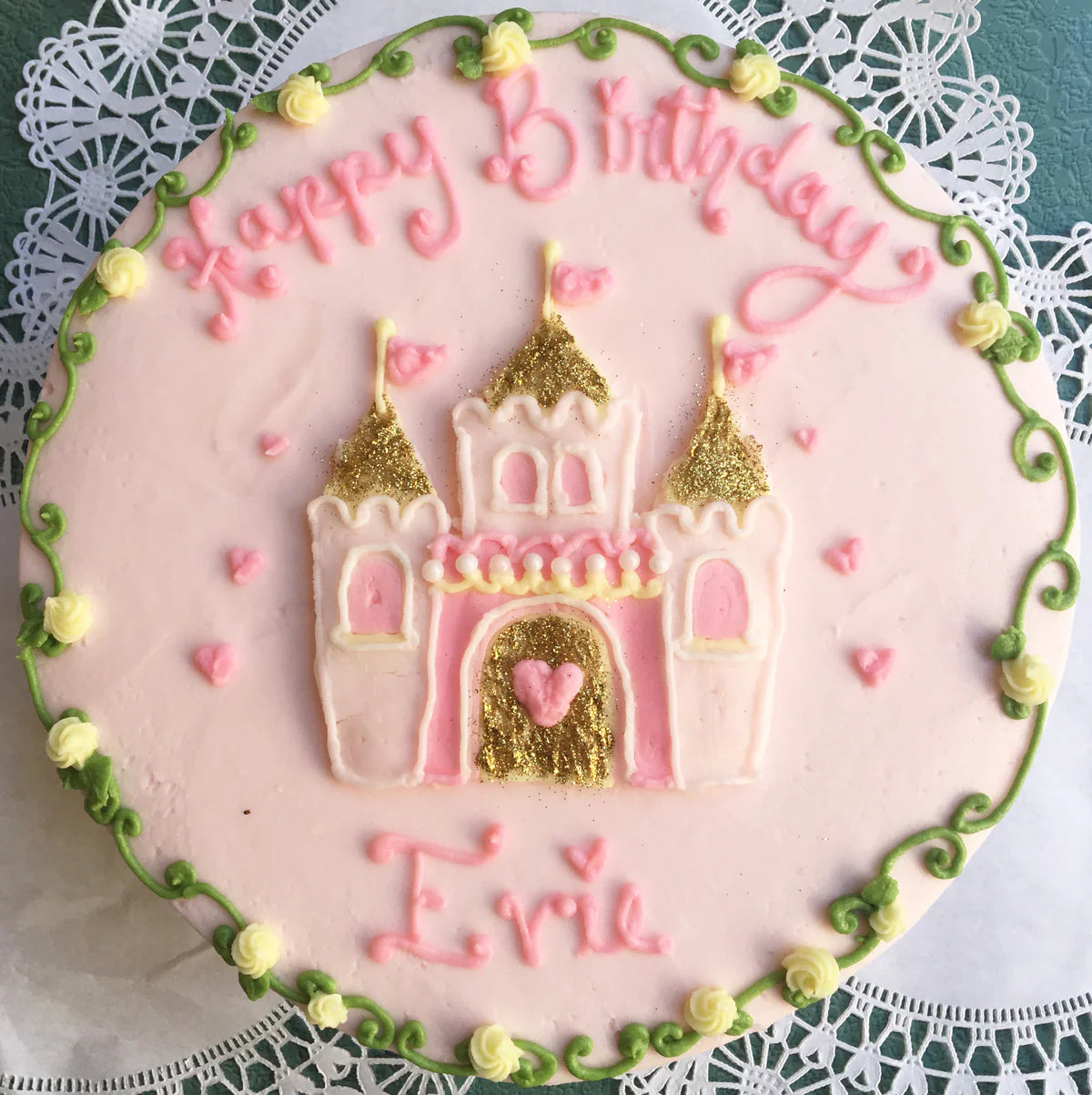 A round pink birthday cake decorated with a pink and gold glitter castle, "Happy Birthday" in pink icing, and "Evie" written below the castle. The cake has a green vine border with yellow roses and small pink heart accents.