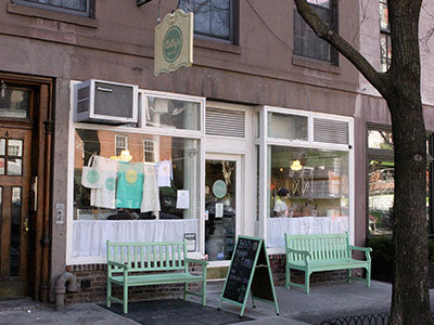 Exterior view of Billy's Bakery in Chelsea, New York City, featuring a brownstone facade, large display windows with merchandise, two green benches, and a chalkboard sign on the sidewalk.