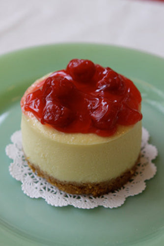 A slice of cheesecake with cherries on top, placed on a plate with a lace-like border.