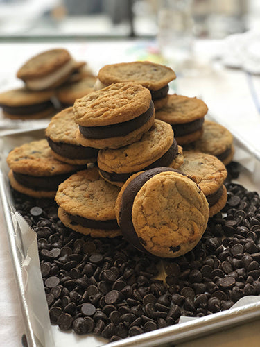A pile of chocolate chip sandwich cookies with dark chocolate or ganache filling, surrounded by loose chocolate chips, on a silver baking sheet.