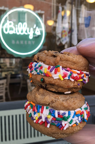 A cookie sandwich with colorful sprinkles on the frosting, held in front of a bakery sign.