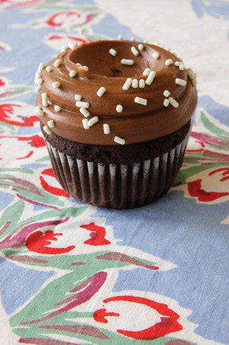 A chocolate cupcake with chocolate frosting and decorative white sprinkles on top, placed on a cupcake holder with a floral patterned fabric underneath.