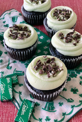 A photo of chocolate mint cupcakes topped with green buttercream and garnished with Andes mint crumble, placed on a cloth with green patterns.