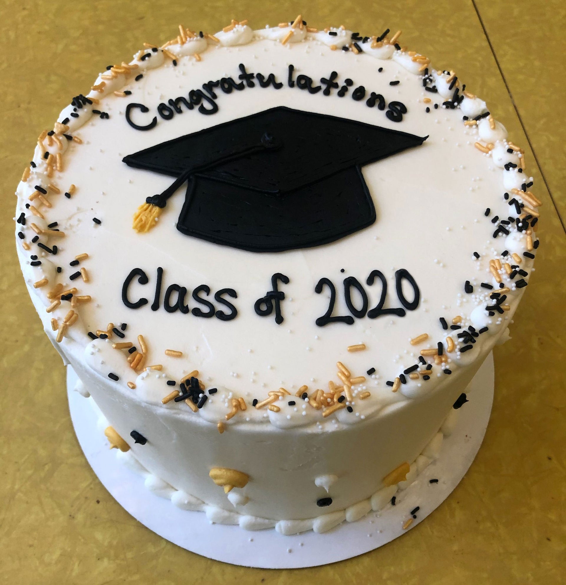 A white round graduation cake on a gold background decorated with black and gold sprinkles, featuring a black graduation cap and tassel. The cake reads 'Congratulations Class of 2020' in black icing.