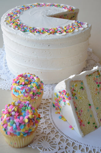 A confetti layer cake with a slice cut out, revealing colorful confetti quins in the cake layers, and two matching cupcakes covered in confetti quins. 