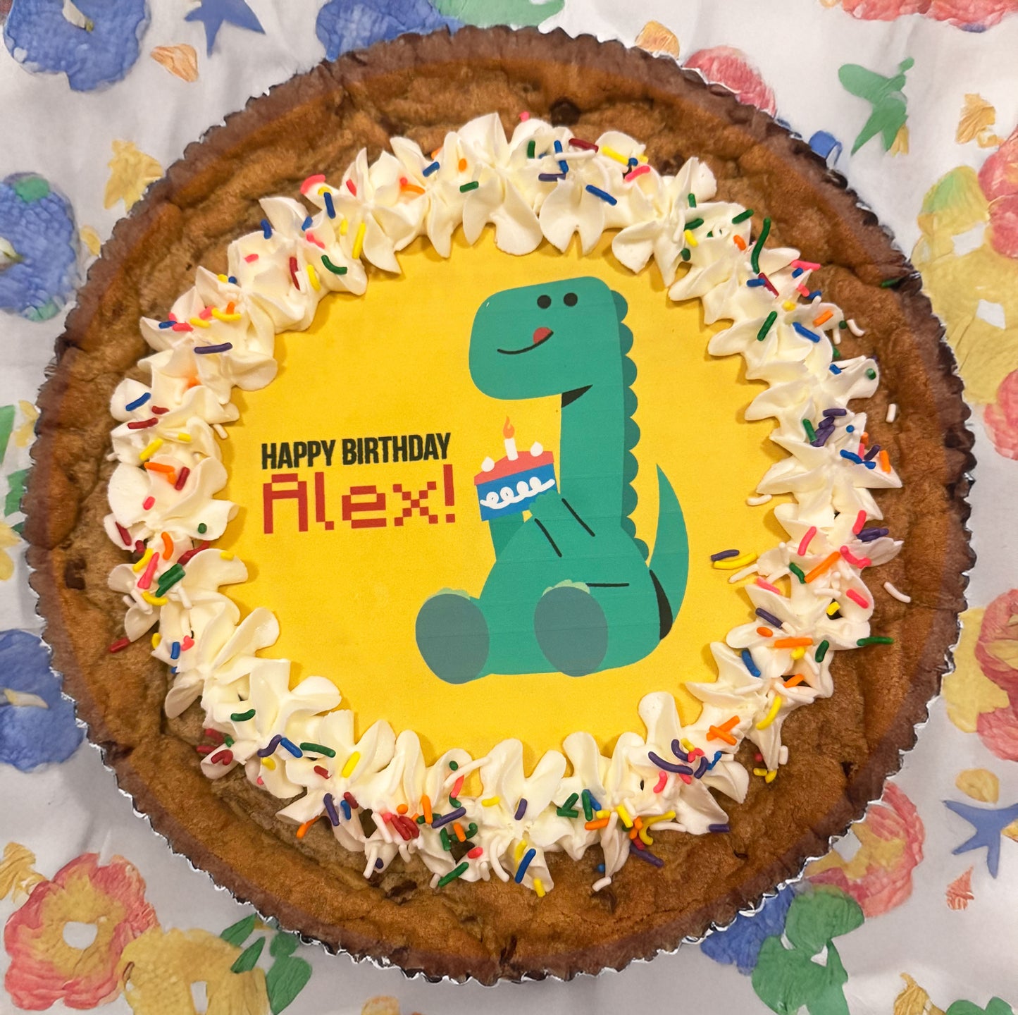 Cookie cake with dinosaur design and 'Happy Birthday Alex!' text on a colorful background