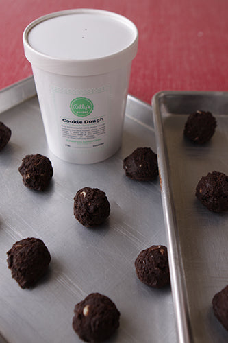 A white plastic tub labeled 'Cookie Dough' with several chocolate cookie dough balls placed on a metal tray in front of the cup.