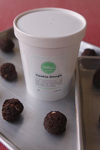 A white plastic tub labeled 'Cookie Dough' with several chocolate cookie dough balls placed on a metal tray in front of the tub.