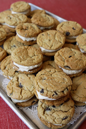 A tray filled with numerous chocolate chip cookie sandwiches, each with a creamy white filling, some resting on white lace doilies.