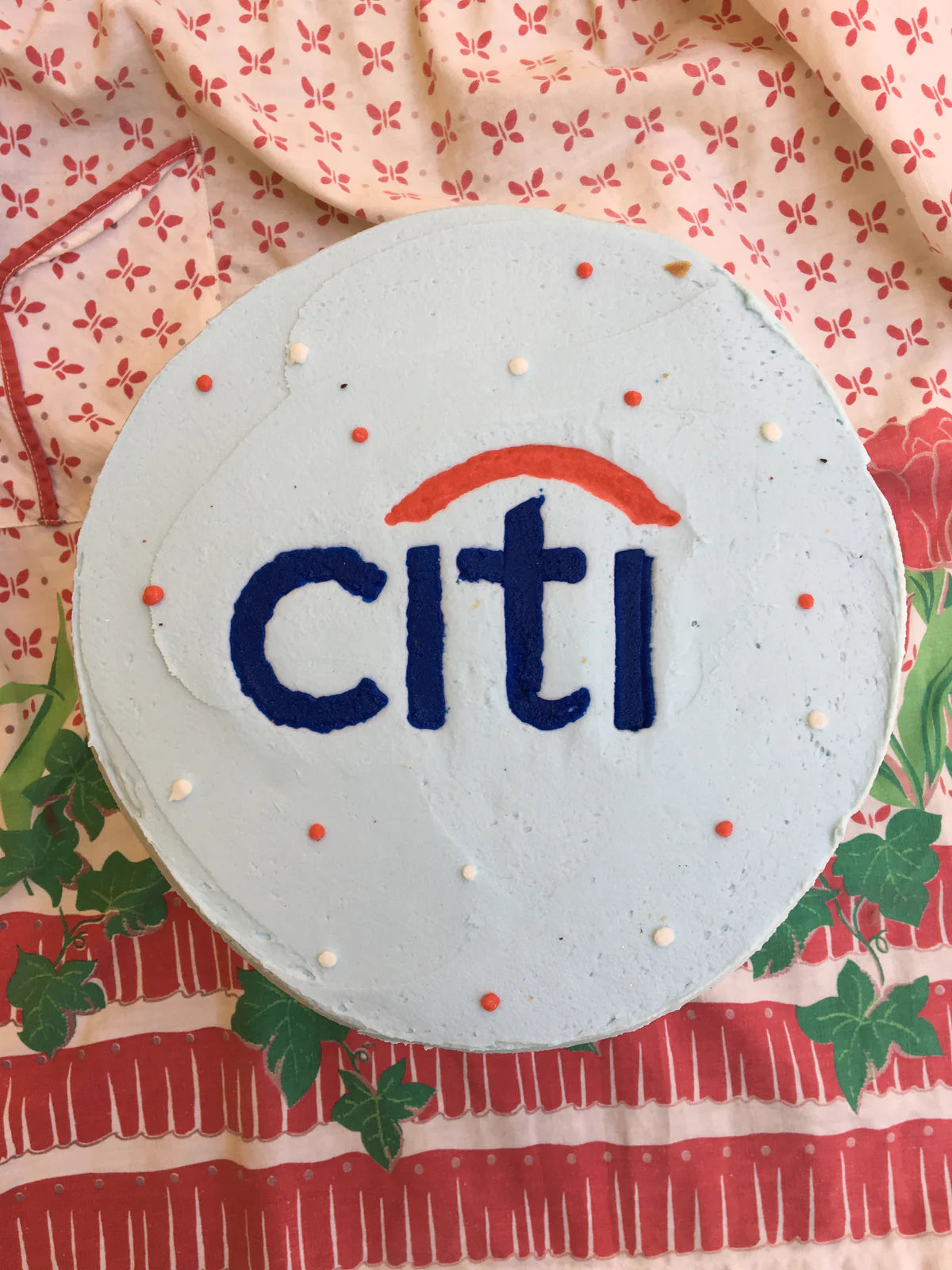 A round cake frosted light blue with the Citi logo in dark blue and orange, surrounded by scattered white and orange sprinkles, resting on a red and white with leaves patterned tablecloth.