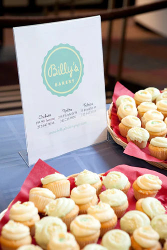Table display featuring a sign for Billy's Bakery with contact information and two baskets filled with frosted cupcakes.