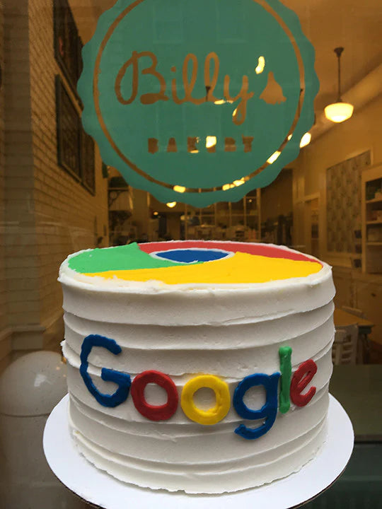 A white frosted cake decorated with the colorful Google Chrome logo on top and the word "Google" spelled out in colorful letters on the side, displayed in front of a bakery window with a "Billy's Bakery" sign.
