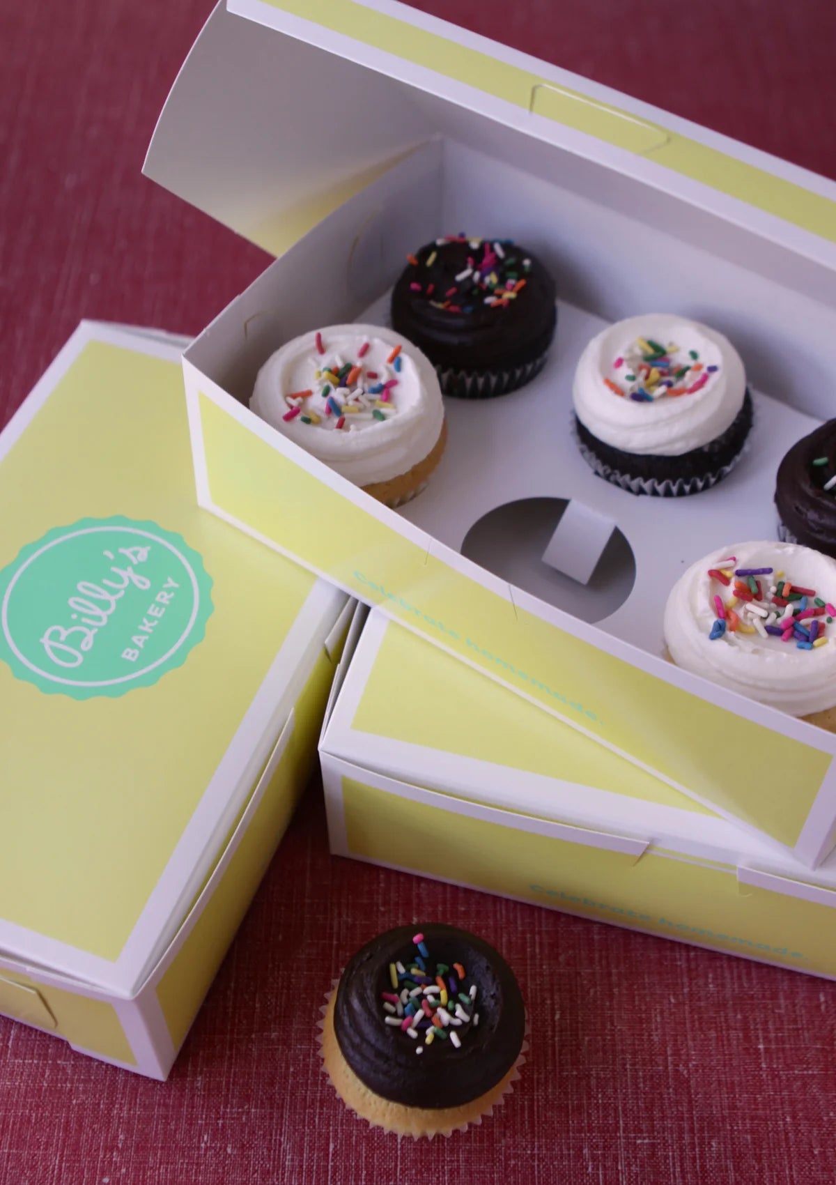 A light yellow box from Billy's Bakery filled with six cupcakes, three topped with white frosting and sprinkles, and three with dark chocolate frosting and sprinkles. One dark chocolate cupcake with sprinkles sits outside the box.
