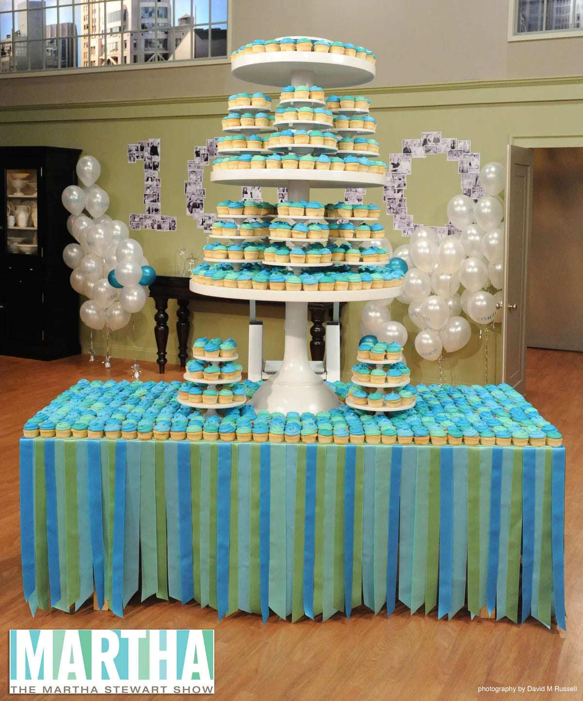 A large, multi-tiered cupcake display with blue and green striped tablecloth, featuring hundreds of teal and yellow cupcakes on a white stand and surrounding table, from The Martha Stewart Show.