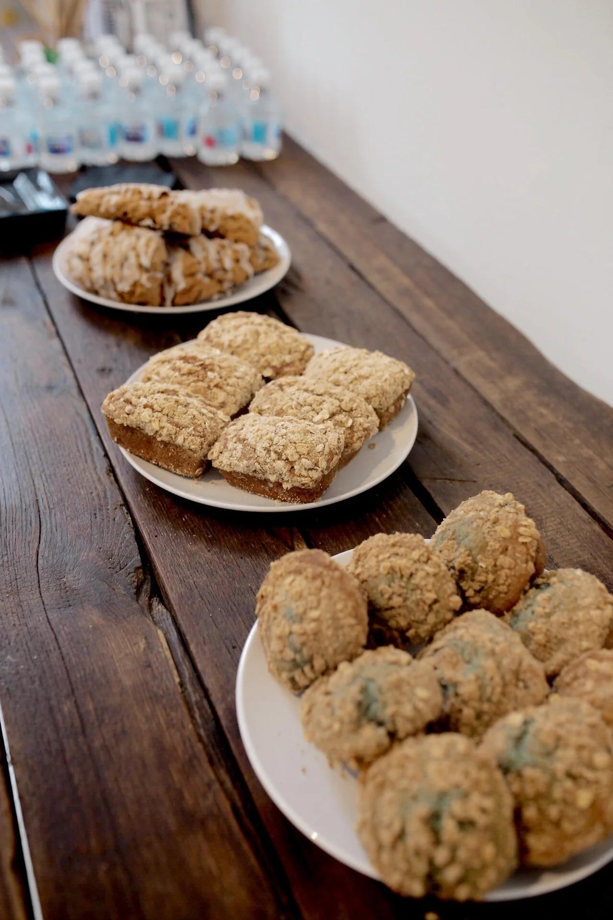 Assortment of baked goods on a wooden table, including muffins with a crumb topping, banana bread and round, textured muffins, with bottles of water in the background.