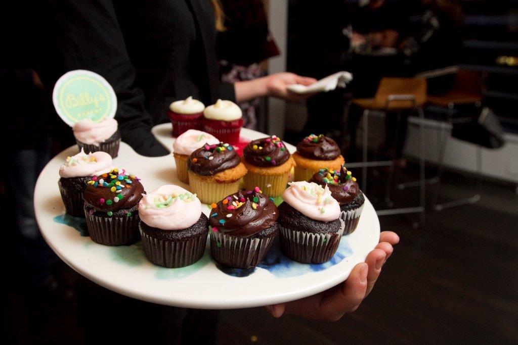 A person holding a white platter filled with an assortment of frosted cupcakes with sprinkles, including chocolate and vanilla flavors, at an event. A small round sign with "Billy's" written on it is visible on the platter.