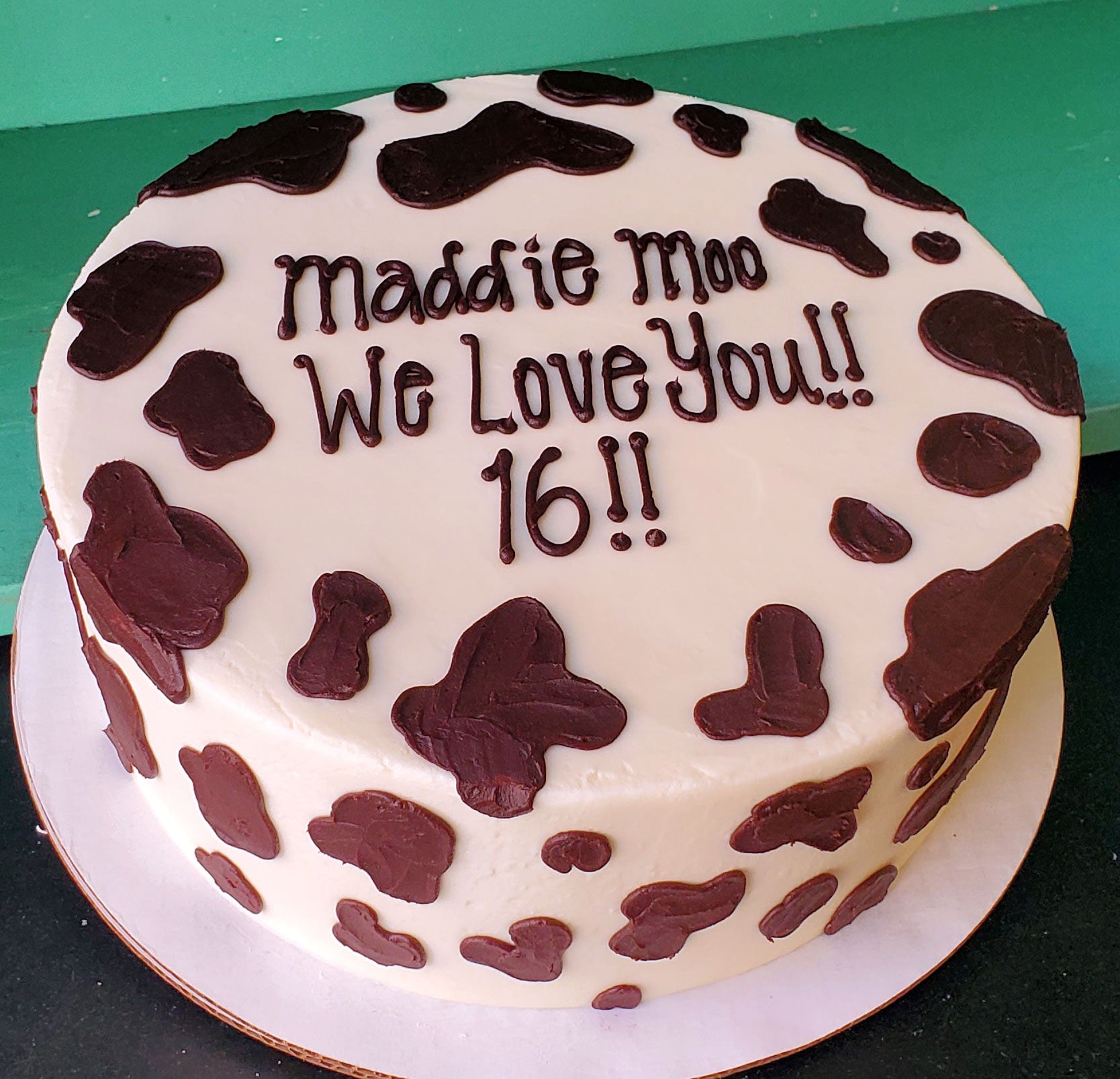 Round white birthday cake with brown cow print spots and chocolate frosting text that reads 'Maddie Moo We Love You!! 16!!'