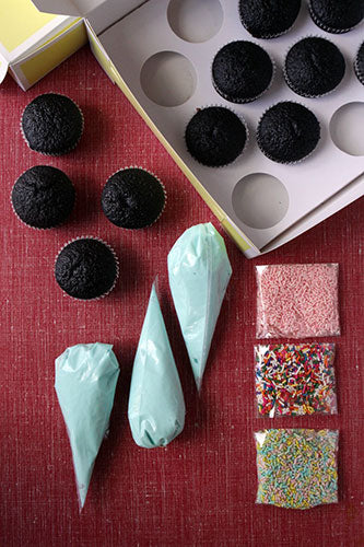 A DIY cupcake decorating kit featuring dark chocolate cupcakes, three piping bags filled with light blue frosting, and three bags of colorful sprinkles (pink, rainbow, and multi-colored) on a red surface. The cupcakes are partially in a white box with empty cupcake slots.