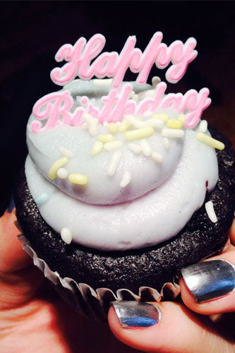 A hand holding a cupcake with a 'Happy Birthday' pick on top, with the text 'HAPPY BIRTHDAY' written in pink frosting.