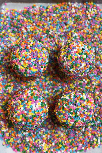 Four round treats covered in colorful sprinkles are arranged on a surface also covered in loose sprinkles, creating a vibrant and textured image.