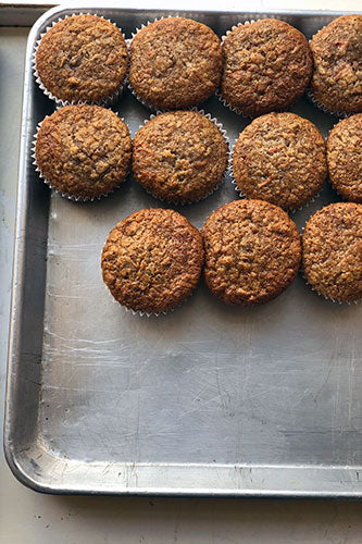 A close-up view of freshly baked, golden brown bran muffins with a textured top, arranged in rows on a silver baking sheet. 